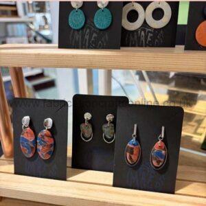 Assorted earrings displayed on black cards on a wooden shelf in a jewelry shop, featuring turquoise circular and orange-blue oval designs.