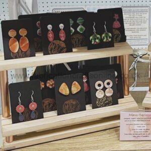 Earrings on black display cards arranged on a wooden rack in a storefront, featuring orange circular and mint-green dangle designs.