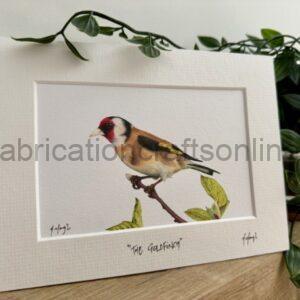 Framed watercolor of a goldfinch perched on a twig, with a white mat and the caption 'The Goldfinch'.