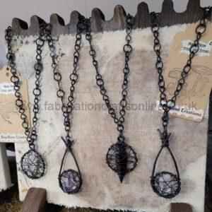 Four handmade wire-wrapped pendant necklaces with dark metal chains, hung on a rustic display; each pendant features a crystal or stone enclosed in an intricate wire cage.