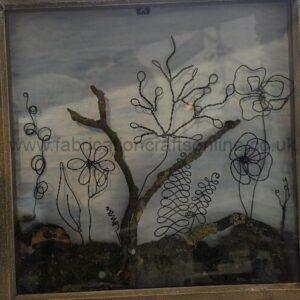 Framed glass display with abstract wire sculptures shaped like trees and flowers rising from a rocky base, set against a pale background.